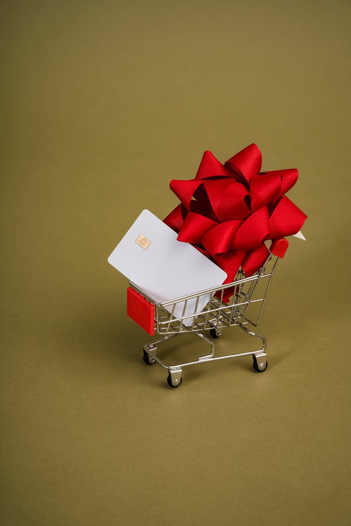 A miniature shopping cart holds a gift card adorned with a red bow, set against a green background.